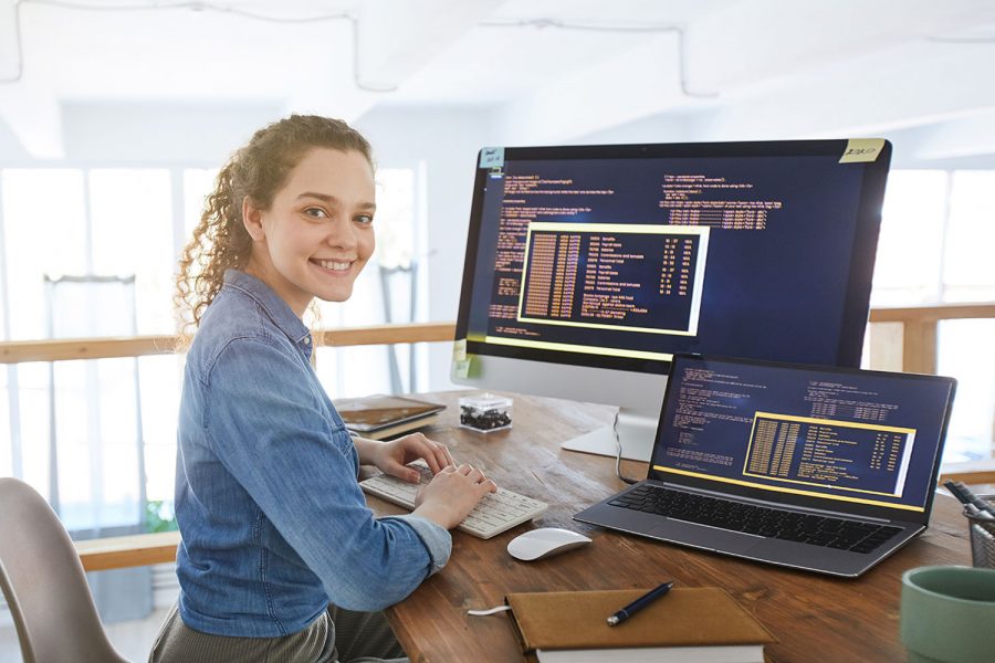 Portrait of female IT developer smiling at camera while typing on keyboard with black and orange programming code on computer screen and laptop in contemporary office interior, copy space