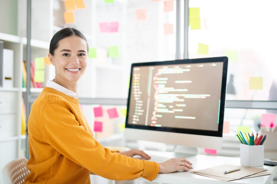 Pretty young webdesigner in casualwear looking at you by her workplace while working over new project