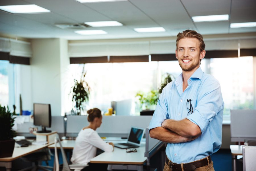 Young handsome successful businessman smiling, posing with crossed arms, office background.